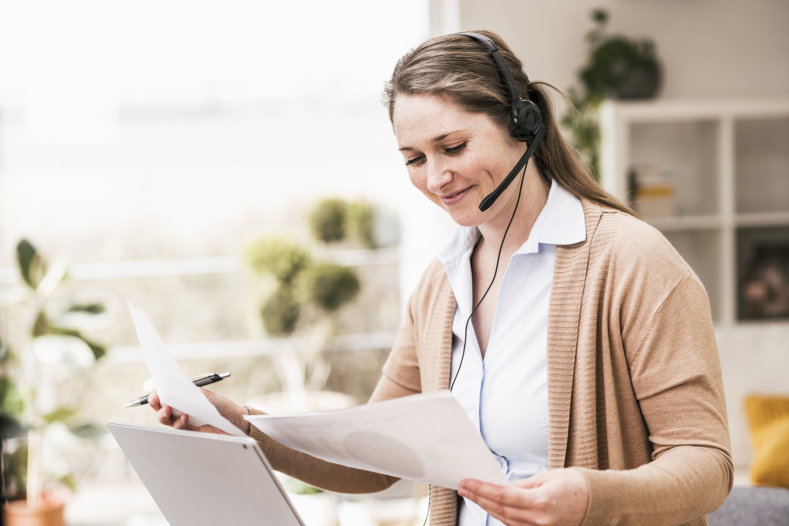 Frau mit Headset vor Laptop hält zwei Papierstücke, symbolisiert Beratung zu Versicherungsverträgen vor Ort oder online