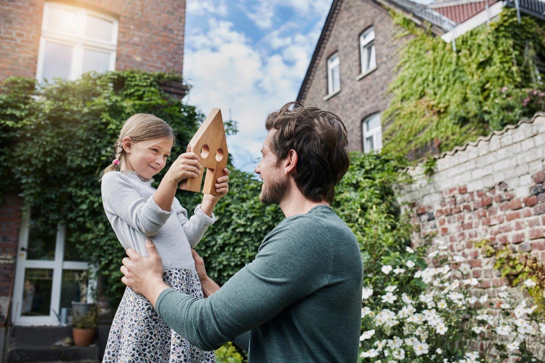 Vater und Tochter im Garten, sie zeigt ihm selbst geschnitztes Holzhaus. uniVersa Wohngebäudeversicherung für das Eigenheim