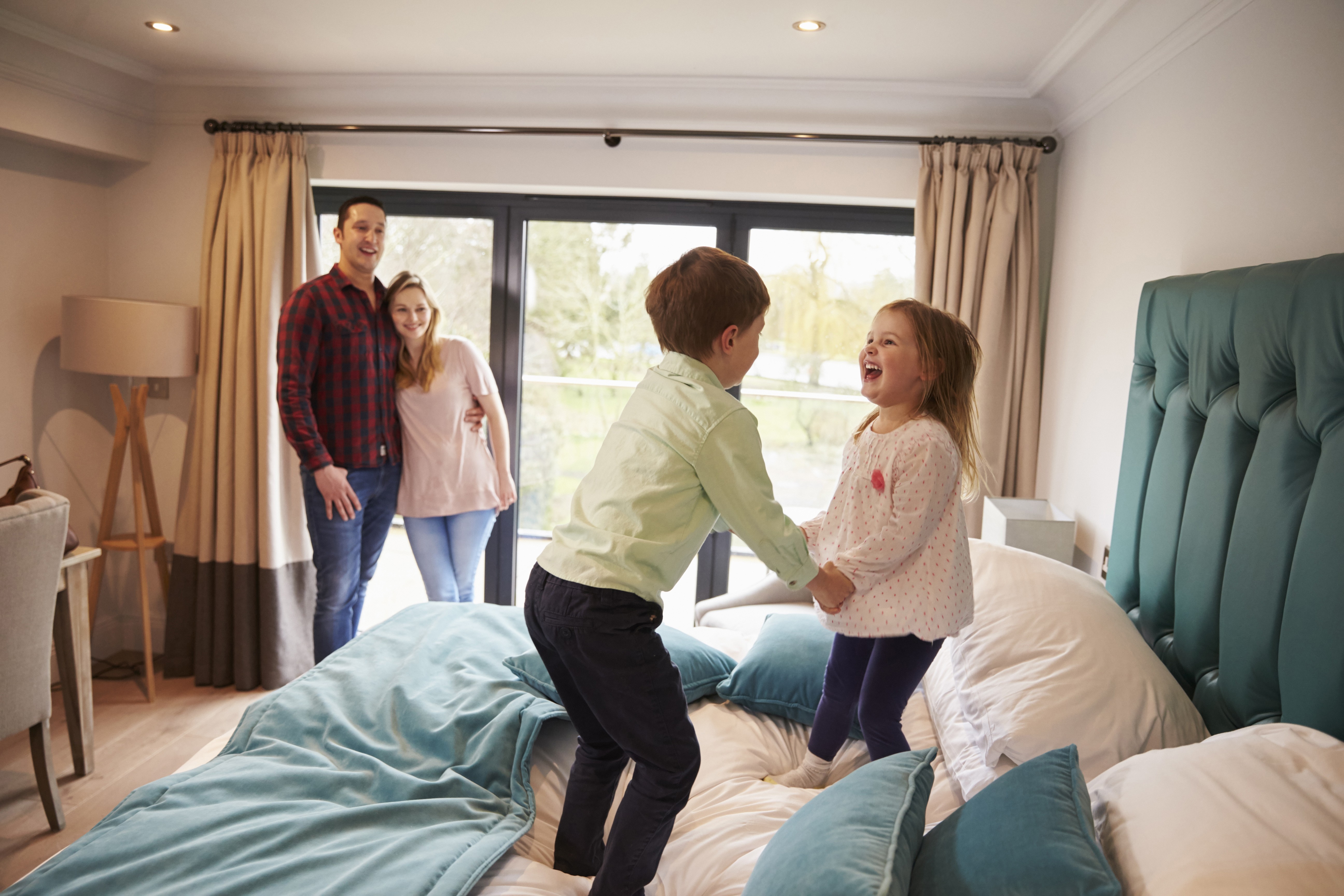 Familie die im Hotelzimmer steht. Kinder springen glücklich auf dem Bett. Symbol für Top Privathaftpflicht für Familien